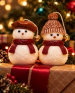 Duas velas de boneco de neve de cachecol vinho apoiadas sobre um presente embrulhado com fita vermelha, diante de uma árvore de Natal iluminada, com pinhas e ramos ao lado